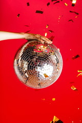 Female hand holds a disco ball on a bright red background and gold glitters fly
