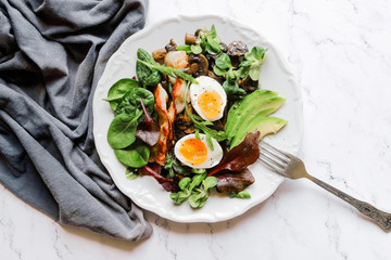 Breakfast eggs, avocado, mushrooms, bacon and fresh arugula greens, spinach on a marble plate dark style top view