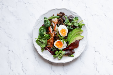 Breakfast eggs, avocado, mushrooms, bacon and fresh arugula greens, spinach on a marble plate dark style top view
