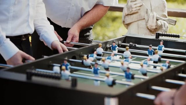 Hands of a man in a white shirt and a man aged with hairy overgrown hands play kicker against girls, actively spinning grips in sunny weather on the street