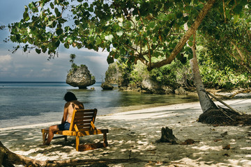Man sitting on his back in a wooden hammock enjoying the beach reading under the shade of a tamarind tree in a Southeast Asian island paradise. Togian islands, Indonesia.