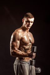 Fototapeta na wymiar Serious tattoed shirtless athlete lifting metal dumbbells training on dark background.