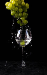 a glass glass stands on the table against a black background. A branch of green grapes hangs from above. Water drops from the top of the grapes into the glass