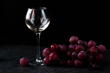 a glass glass stands on the table against a black background. There are red grapes nearby. On top of the glass is poured wine from a bottle with a dark glass