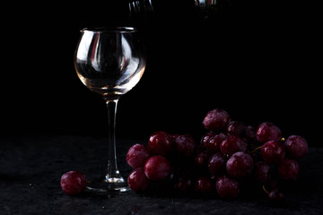 a glass glass stands on the table against a black background. There are red grapes nearby. On top of the glass is poured wine from a bottle with a dark glass