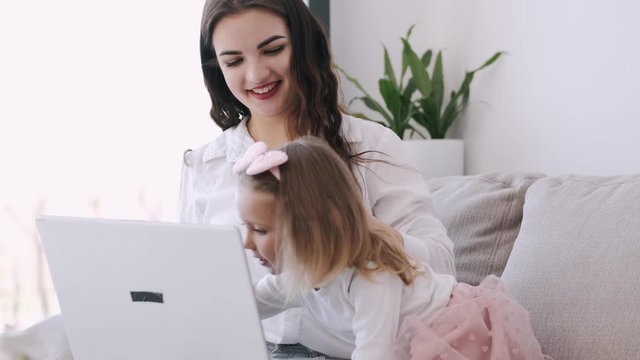 The pretty mother and her child girl are studying with a laptop. The mother is teaching heer kid. The girl is getting along with the parent typing on the keyboard and watching something on hte laptop