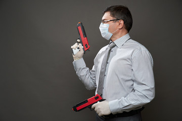a man dressed as a businessman, posing in studio on gray background, medical face mask and protective gloves, gun, glasses, shirt and tie - concept of quarantine and antivirus protection