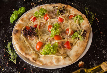 Pizza with Caesar dressing, meat, grated cheese, chopped tomatoes and salad. On a black table with sprinkled with spices and ingredients. Photo for the menu.