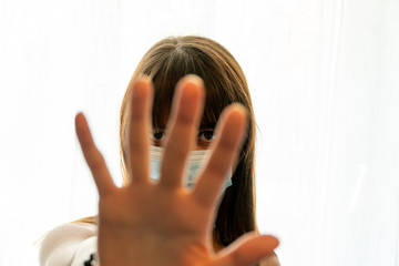 Woman with facemask peeking through fingers signaling to stop with hand on white background