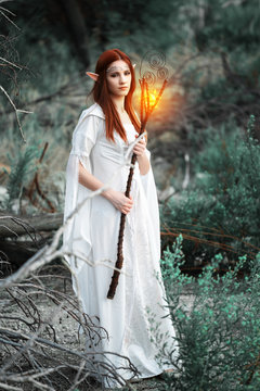 
a girl with elven ears in a white dress walks through the woods and holds in her hands a magic staff, glowing