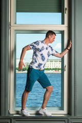 Tall handsome man wearing white t-shirt and blue shorts standing by big window in interior