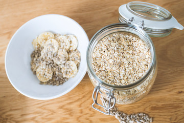 A glass with oatmeal and a bowl with banana, oat and sunflower seeds on wooden background