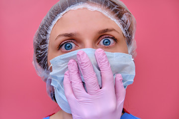 Portrait of a doctor woman with a fright in the eyes, closeup. Nurse in a blue uniform and a protective mask on a pink background.