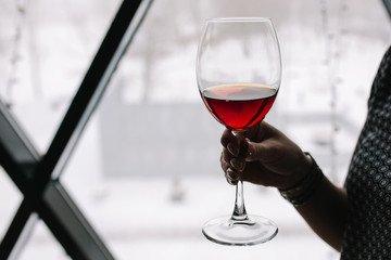 a glass of  red wine in a female hand on a window background. drink red wine in good company. say a toast at the table. hold a glass with wine in hand. a glass of wine for communication.