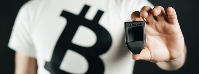 A man dressed in a white shirt with logo Bitcoin is holding a crypto hardware wallet - Trezor in his hand on the black background banner.