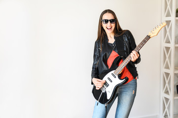 A young beautiful woman in sunglasses and in leather jacket is playing an electric guitar indoors,...