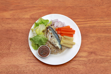 Breakfast in the white round plate on wooden floor. Fried mackerel and Rice with Spicy Shrimp Paste Dip and vegetable, Chinese cabbage, baby corn, carrots and sliced onions.