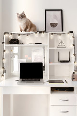 White home office interior, cozy light workplace with laptop and garland cat sitting on the shelf