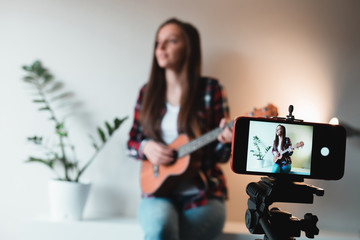 Girl in a shirt and jeans writes a vlog on the phone about playing ukulele.