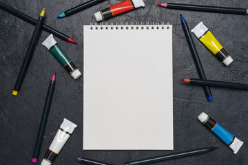 Artist's desk flatlay with a sketchbook, paints and watercolor brushes on a dark background