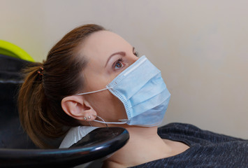 A young woman in a medical mask washes her hair in a barbershop during the covid-19 coronavirus...