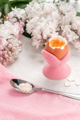 Soft-boiled egg on a pink stand with a spoon and lilac flowers on a light background