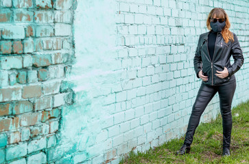 A young red-haired girl in sunglasses and a black medical mask walks the streets of the city during the coronavirus epidemic. Girl in black on a background of a brick wall. COVID-19