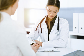 Woman-doctor and patient sitting and talking at sunny hospital office. Green color blouse suits to therapist. Medicine concept