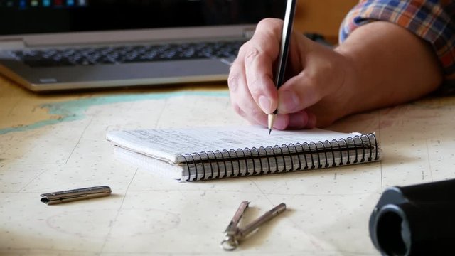 The man traveler is writing in a notebook with a pencil. Closeup of man hand, paper notebook placed on the sea map, and other navigator tools. Home workplace for voyager. Preparing for travel