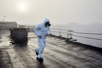 Man wearing white coverall and gas mask, running on the roof of an urban building with his back to...