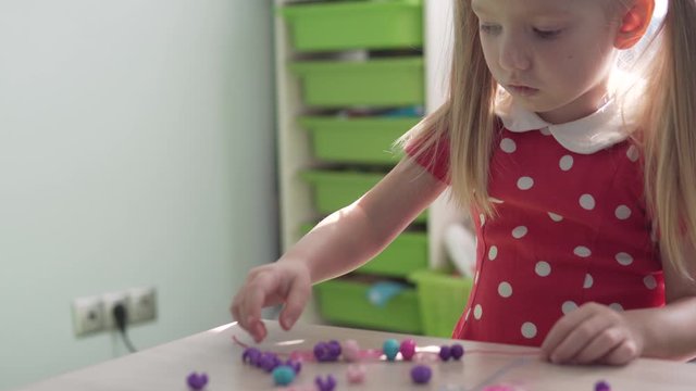 A little girl makes a bracelet out of beads. The development of fine motor skills in children at home.