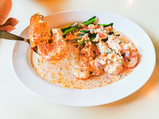 Shrimp in a creamy sauce with vegetables on a white plate With the hand of a woman using a spoon to scoop up 1 shrimp