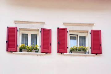 Two Italian windows on the white wall facade with open red color classic shutters and flowers on the windowsill