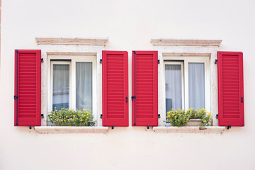 Two Italian windows on the white wall facade with open red color classic shutters and flowers on the windowsill