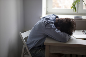 A tired man in a blue office shirt laid his head on his hands at a table opposite the window.