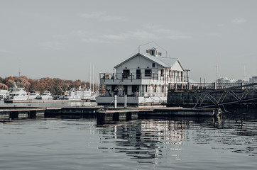 Fototapeta na wymiar An aesthetic calm view of an authentic boathouse on a river pier. White wooden building with a parked yachts. Reflection on the water. Cloudy sky. Travel and sailing concept. River landscape