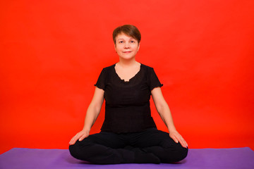 Fototapeta premium Aged woman in black clothes doing yoga sitting in lotus position on a purple rug. Studio photo on a red background. 