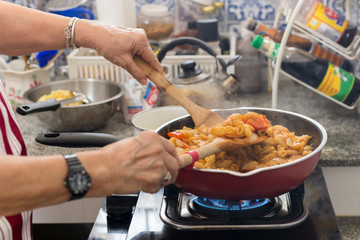 Old woman cooking macaroni pasta in flying pan in the kitchen.