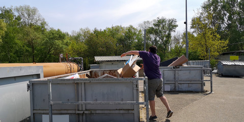 Plump,dark-haired,middle-aged man throws cardboard box with household waste into huge container at station of collection of large-size garbage in Germany, Europe on sunny day.Selective focus.Mockup.