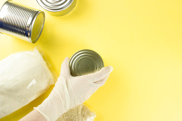 Food for quarantine isolation period flat lay on yellow background with copy space. Sugar, rice, female hand in glove taking canned food. Crisis food supplies. Delivery, donation concept. Stock photo.