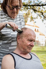 Woman cutting hair with electric razor to senior man outdoors at garden. Elderly people take care...