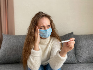girl in a disposable medical mask is sitting at home with a headache. The girl has a high temperature.