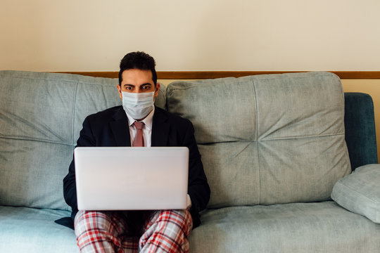 Executive Businessman With A Mask On His Face Working From Home With His Laptop. Dressed In A Suit And Tie And Pajama Pants. Sitting On The Couch. Concept Work From Home. Coronavirus Pandemic.