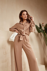 Beautiful brunette woman natural makeup wear fashion clothes casual dress code office style total beige blouse and pants suit, romantic date business meeting accessory bag interior stairs flowerpot. - obrazy, fototapety, plakaty