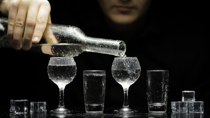Barman pour frozen vodka from bottle into glasses with ice. Black background