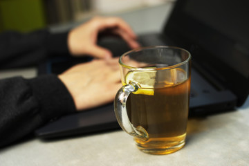 Young woman working on a laptop on the kitchen table, remote work at home, self-isolation, selective focus on a Cup of tea with lemon