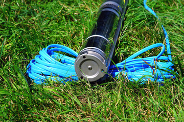The bottom of the pump with cable on a background of green grass. The bottom cover of the borehole pump. Electric pump with blue cable. - obrazy, fototapety, plakaty