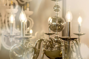 The picture of a glowing chandelier with metal chrome fixtures and shades in the form of candles. The concept of the classical style in the interior. Close up. Soft warm filter, selective focus.