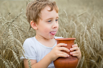 a small boy in a wheat field drinks milk from a brown ceramic jug