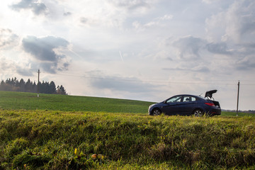 Fototapeta premium Car standing in the fields. Sky and clouds in background. Forest, trees on a hill. Green grass, yellow flowers. 
